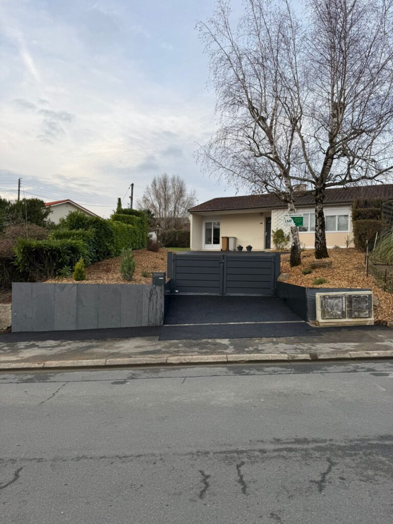 Accès véhicules terminé avec muret de retenue en ardoise et portail moderne, devant une maison après travaux paysagers.